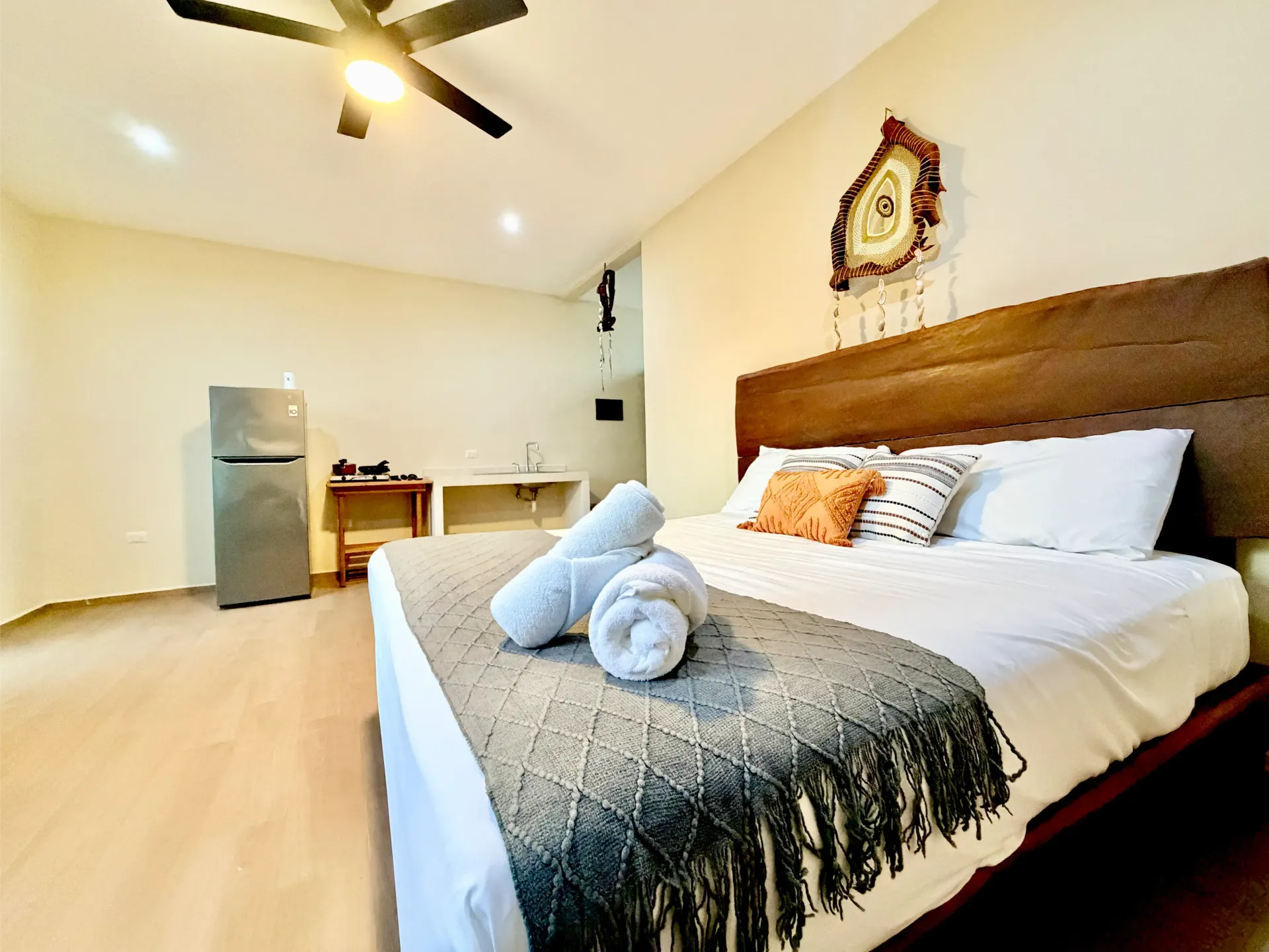Bright bedroom with a large bed, decorative towels, ceiling fan, and small desk near a mini fridge.