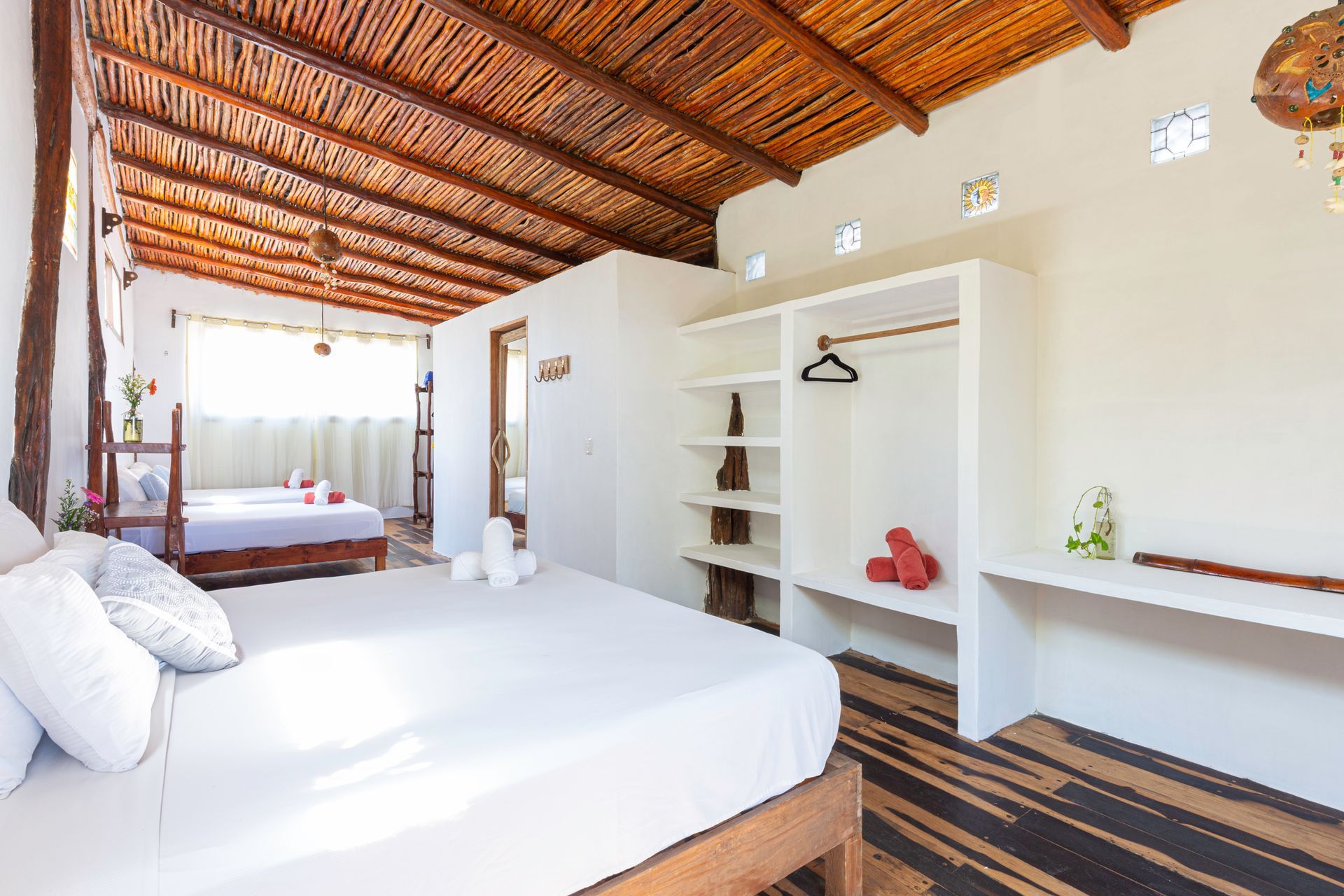 Bright bedroom with a wooden thatched ceiling, white bed, open closet, and a second bed in the back.