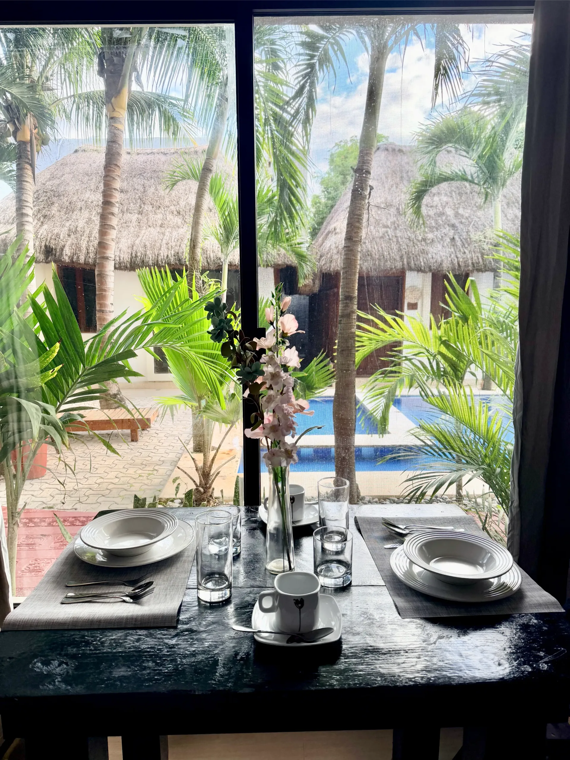 Dining table by a window overlooking a tropical patio and pool, with place settings and a vase centerpiece