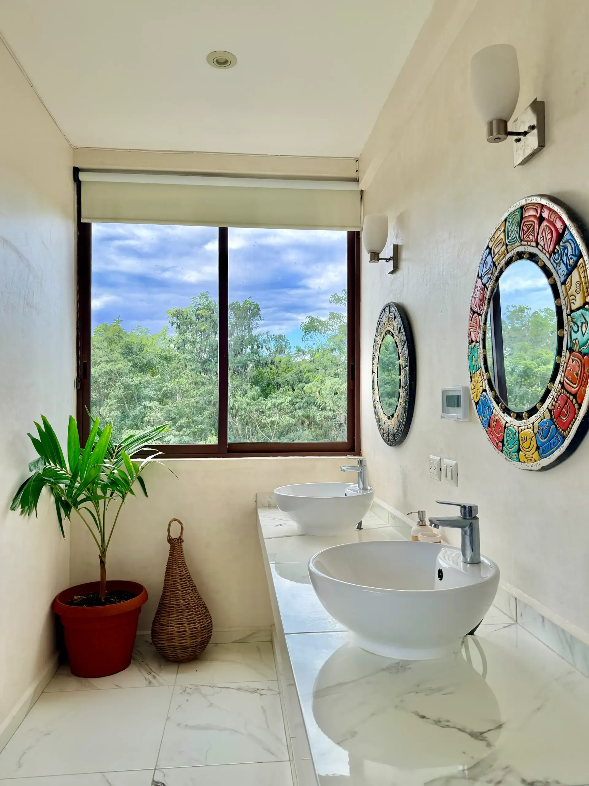 Bright bathroom with vessel sink, round colorful mirror, plants, and a large window overlooking greenery