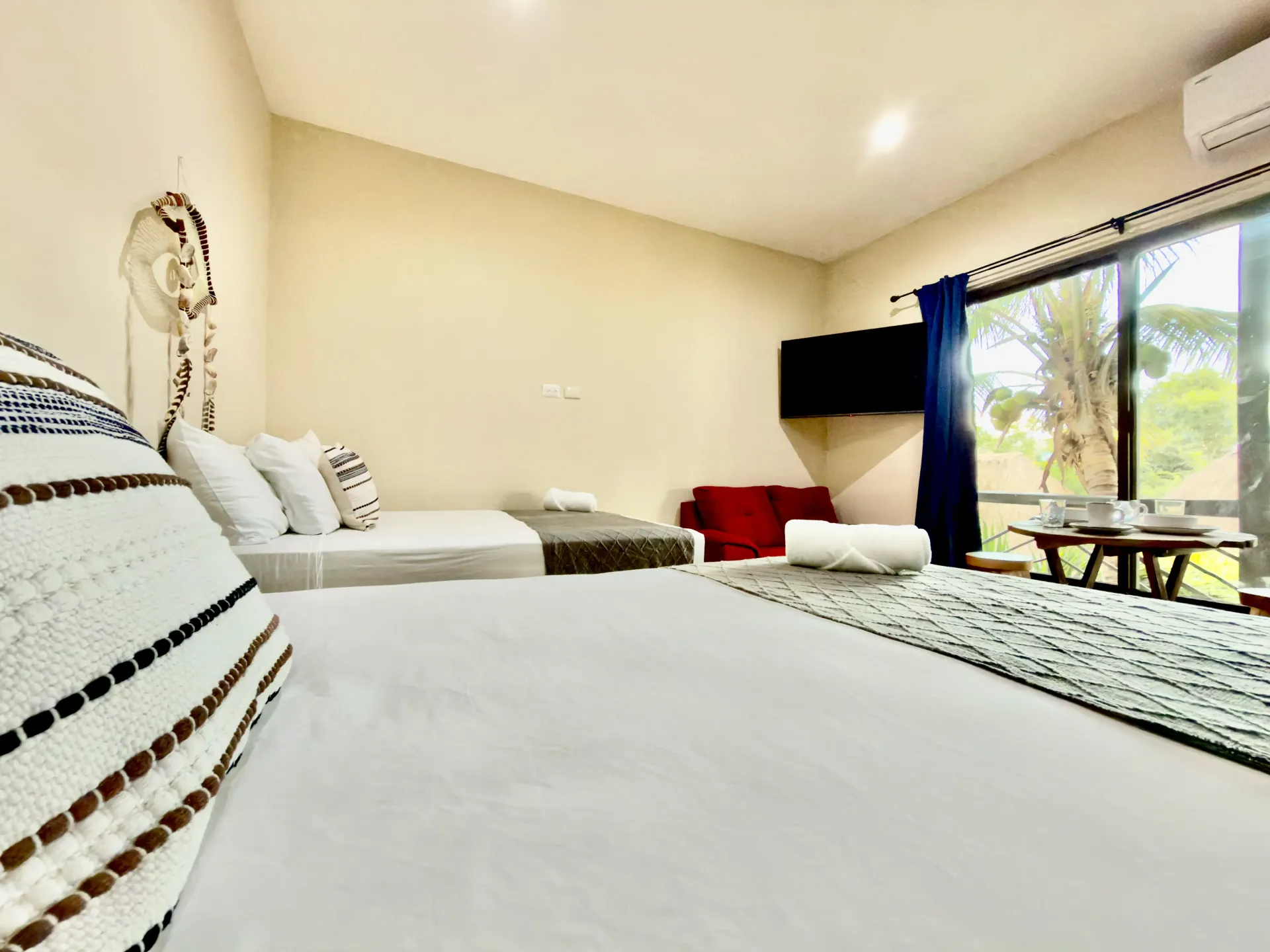 Bright hotel room with two beds, striped pillows, a TV, and a large window with ocean view.