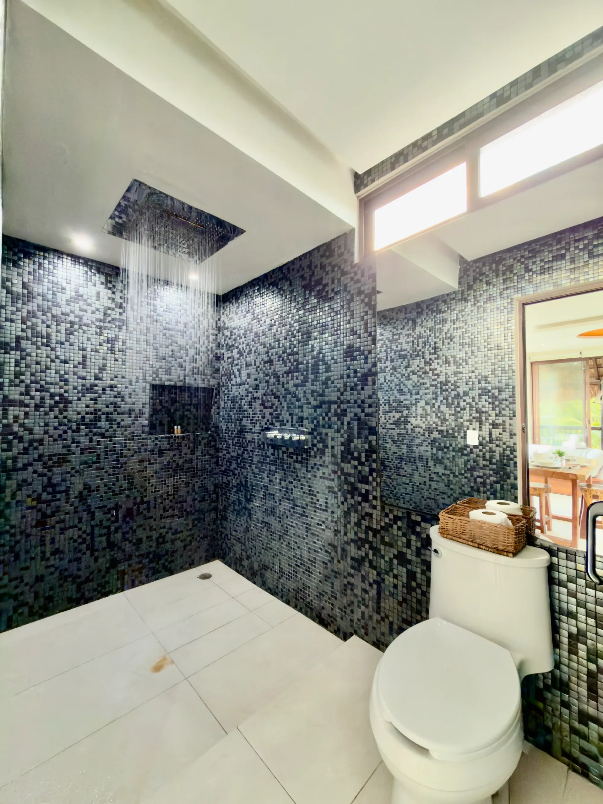 Modern bathroom with dark mosaic tile shower, white floor, toilet, and window light