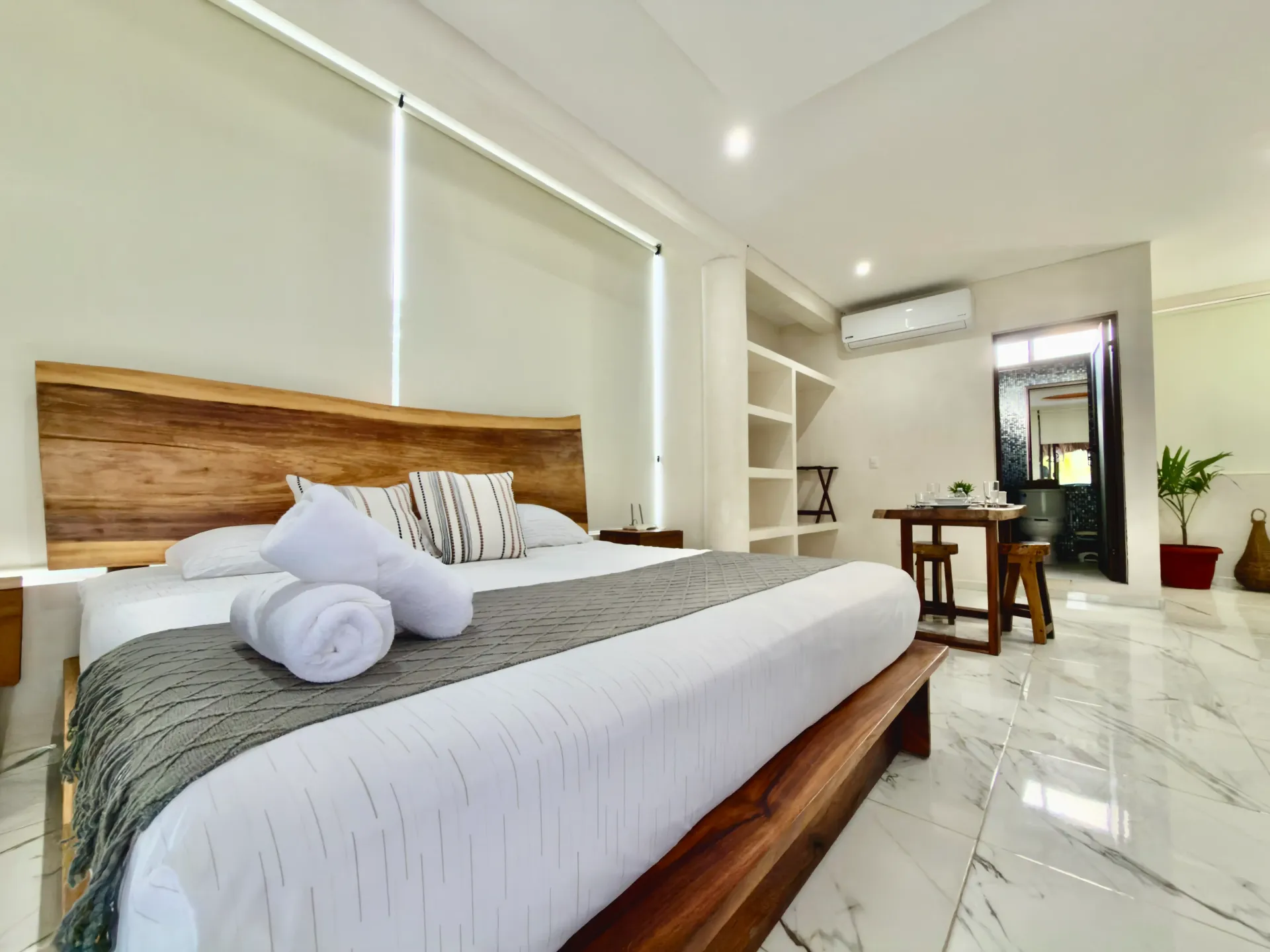 Bright bedroom with a wooden bed, white linens, shelves, desk, and balcony doors in a modern apartment