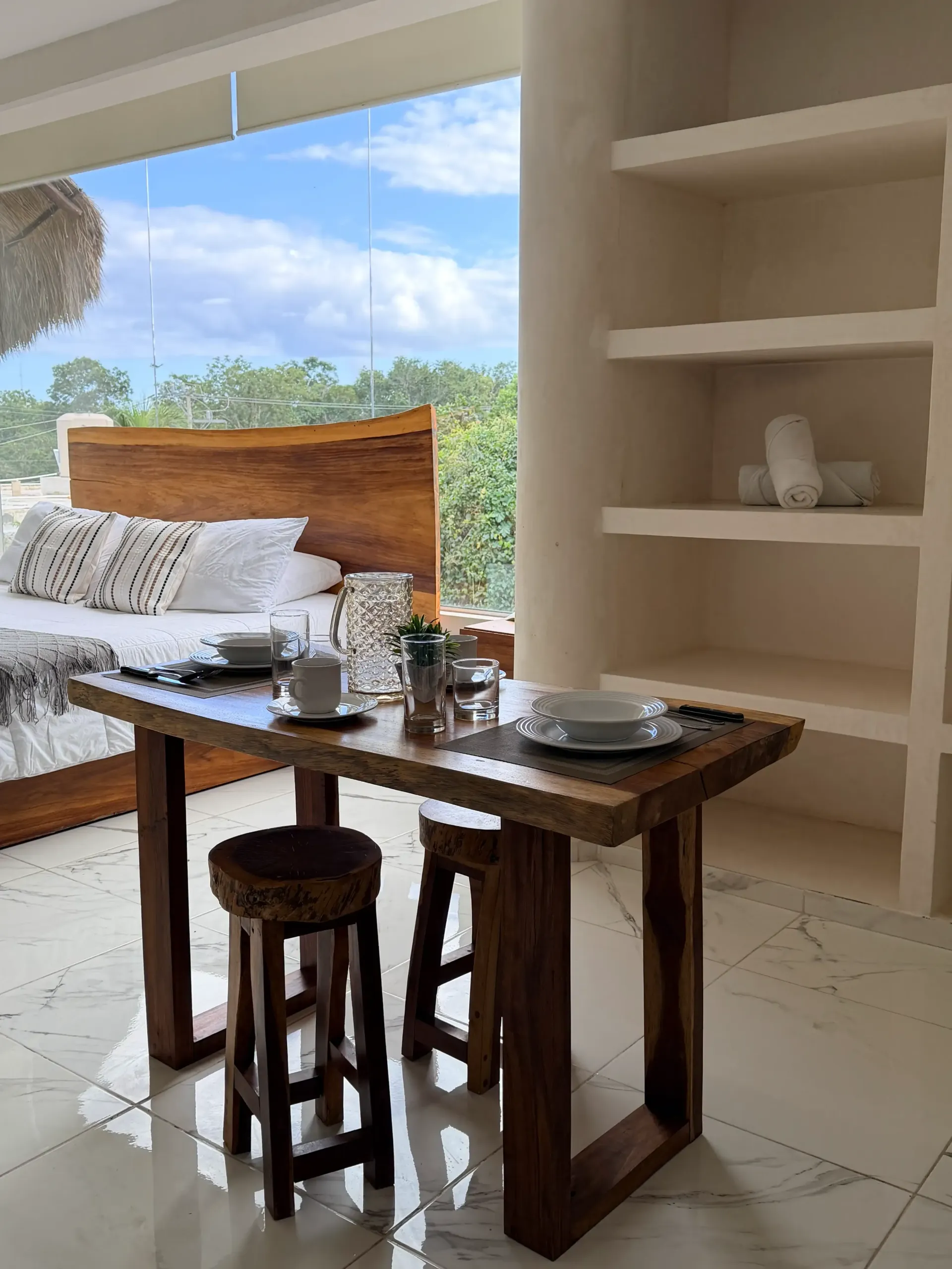 Cozy open-air dining nook with wooden table, stools, built-in shelves, and a bench by a bright window view