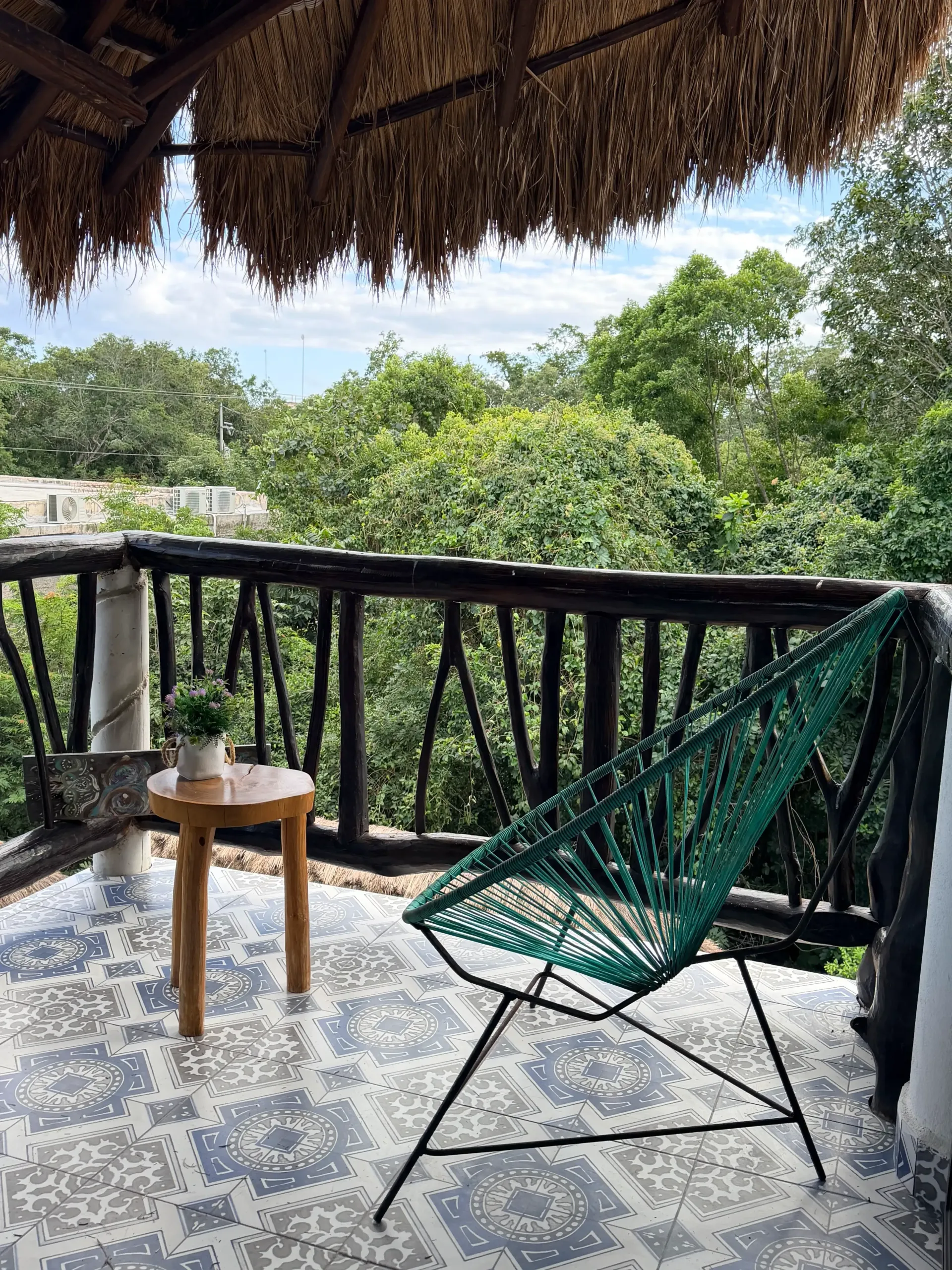 Thatched balcony with black railing, green chair, and small wooden table overlooking trees