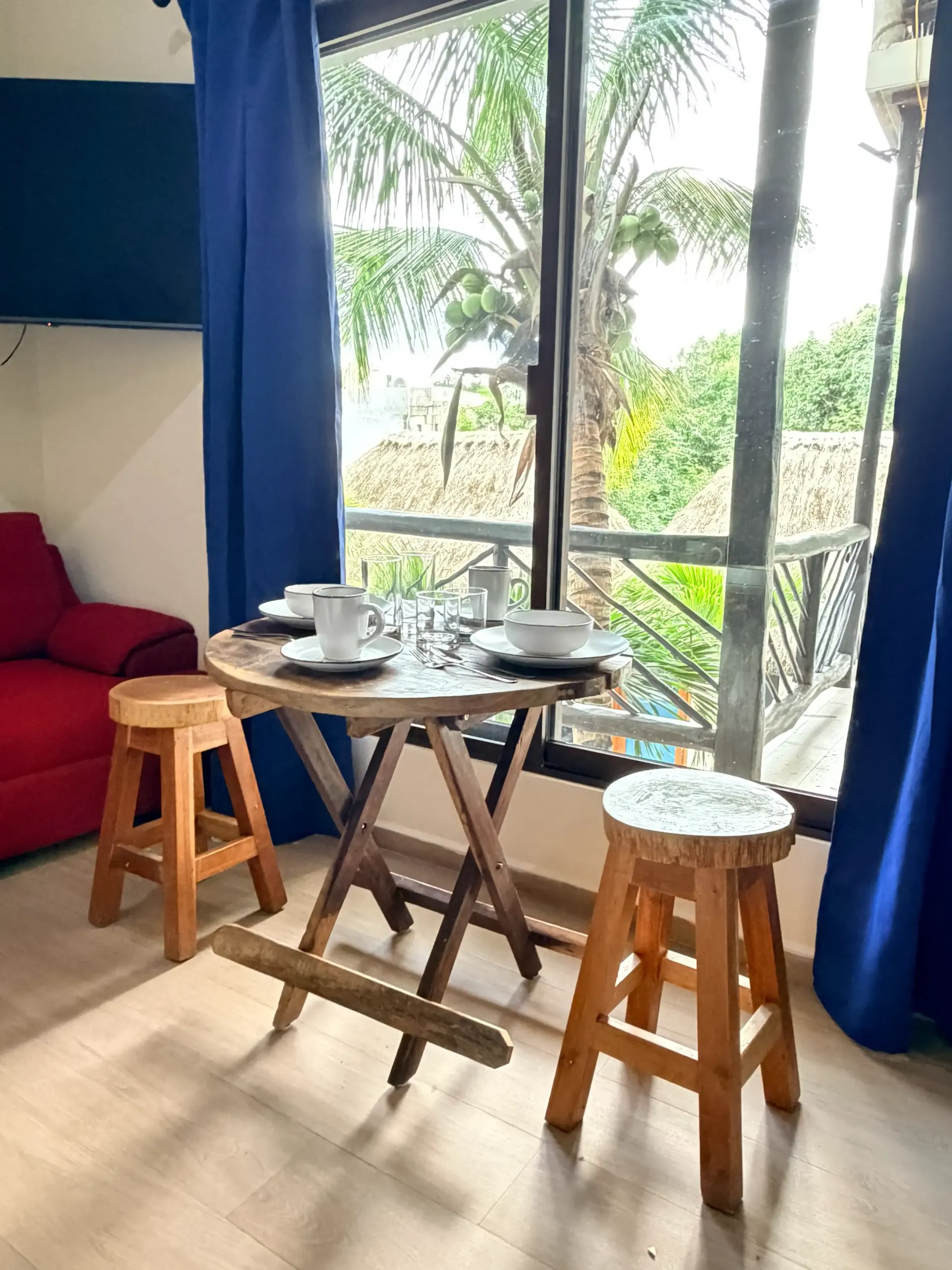 Small table by a window with two stools, dishes, and blue curtains in a bright room