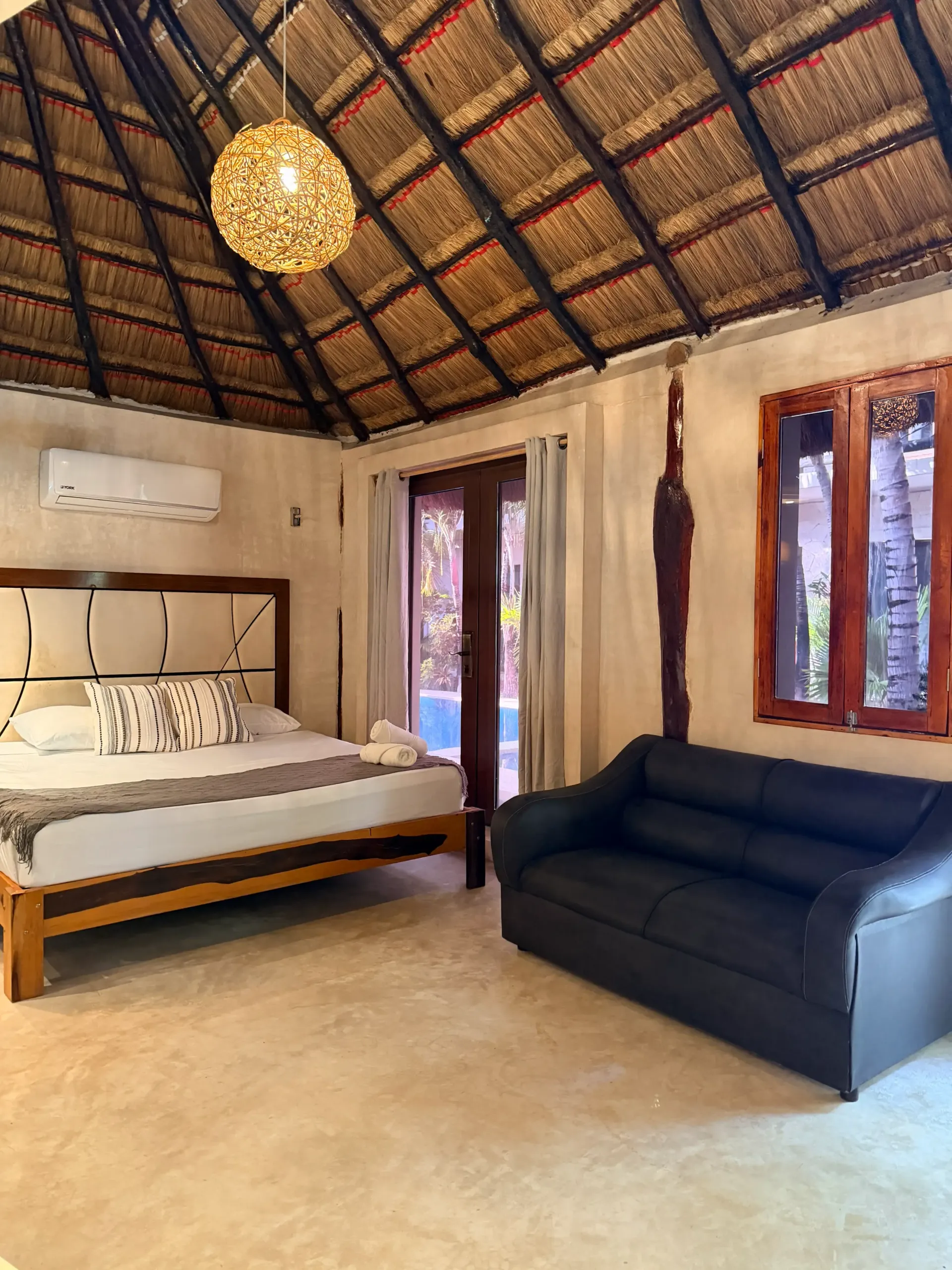 Rustic bedroom with thatched ceiling, wooden bed, dark sofa, and French doors to a patio.