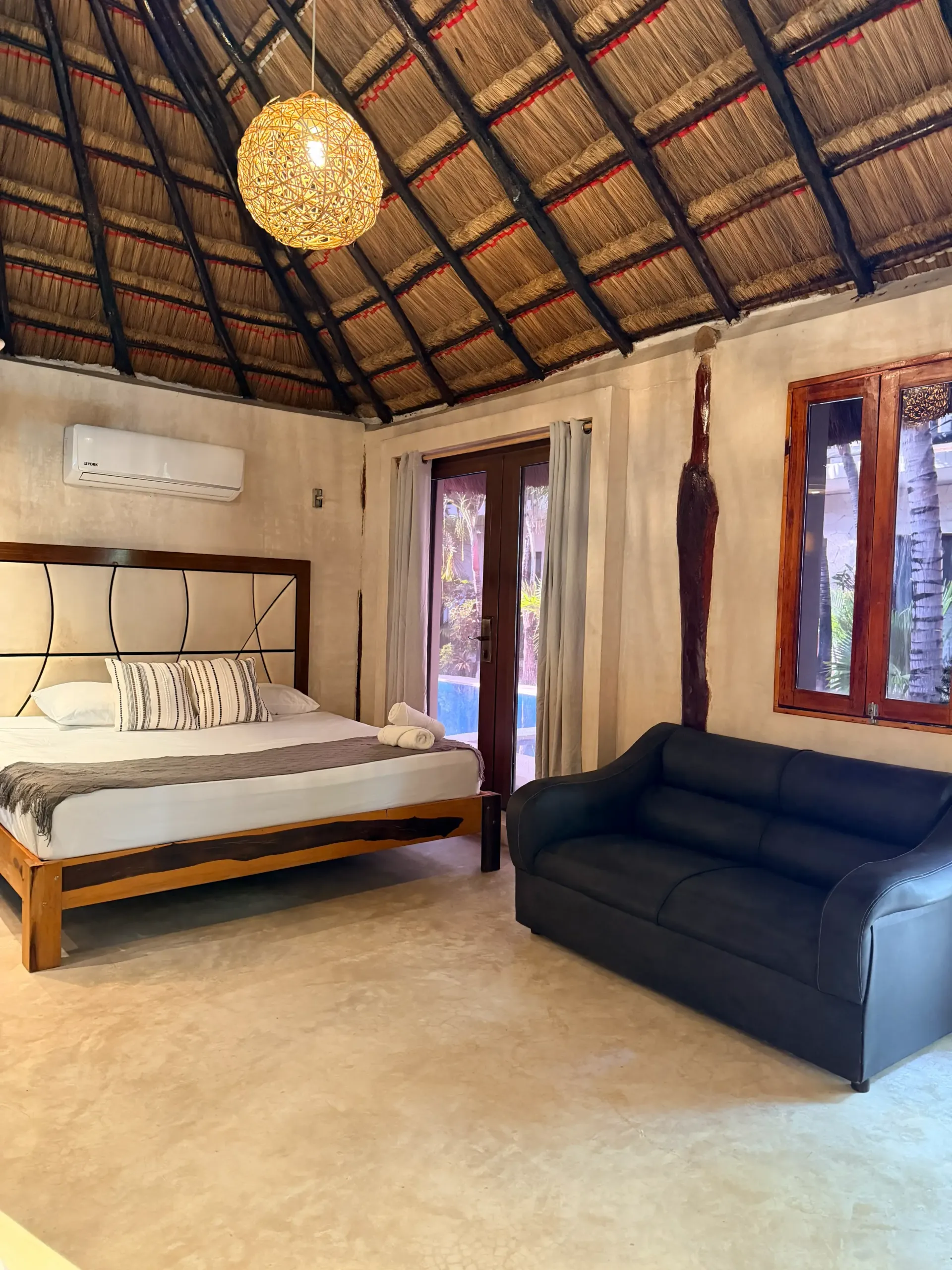 Rustic bedroom with thatched ceiling, four-poster bed, black sofa, and tall windows.