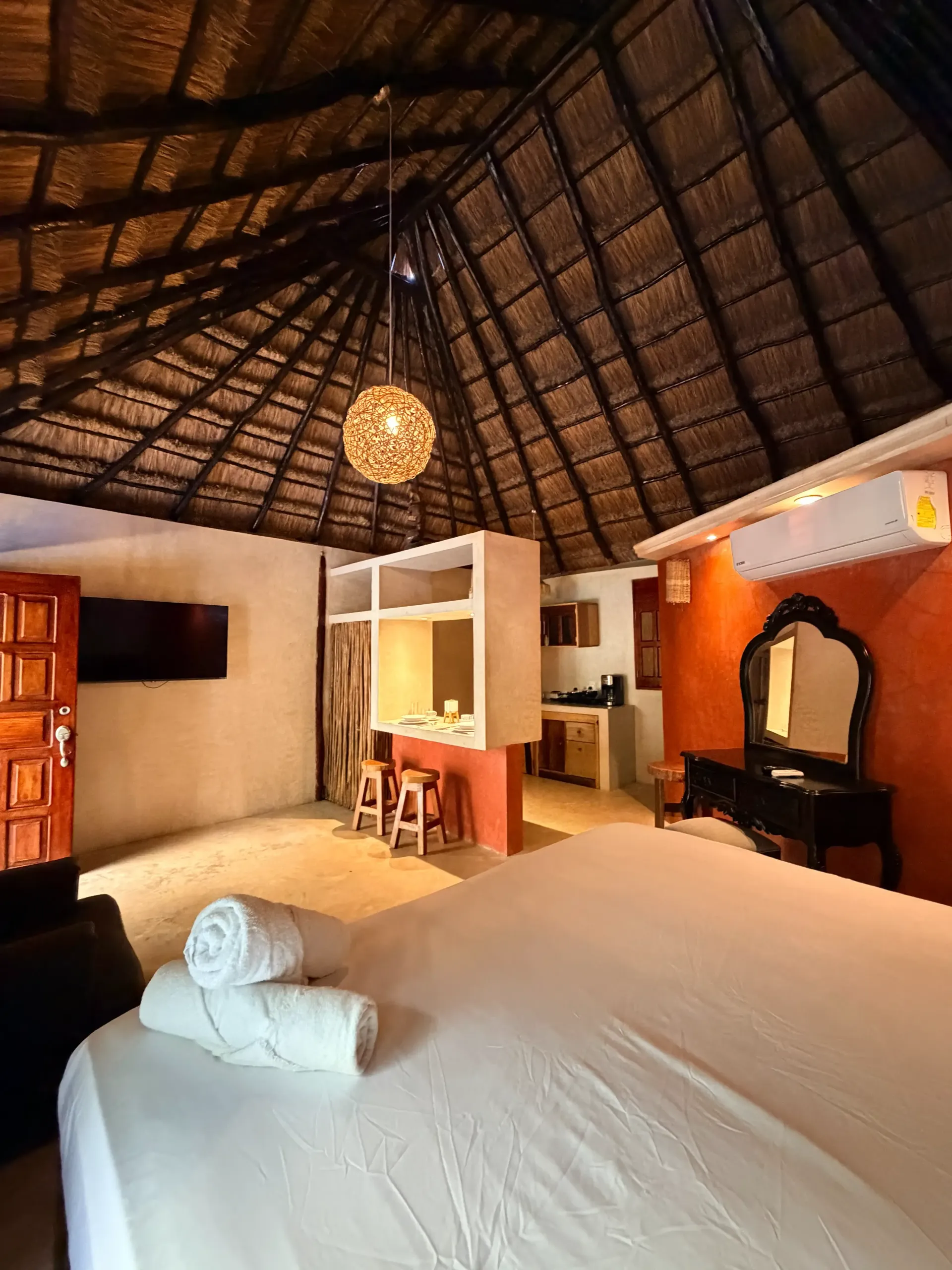 Thatched-roof bedroom with a large bed, hanging light, and vanity in warm tones