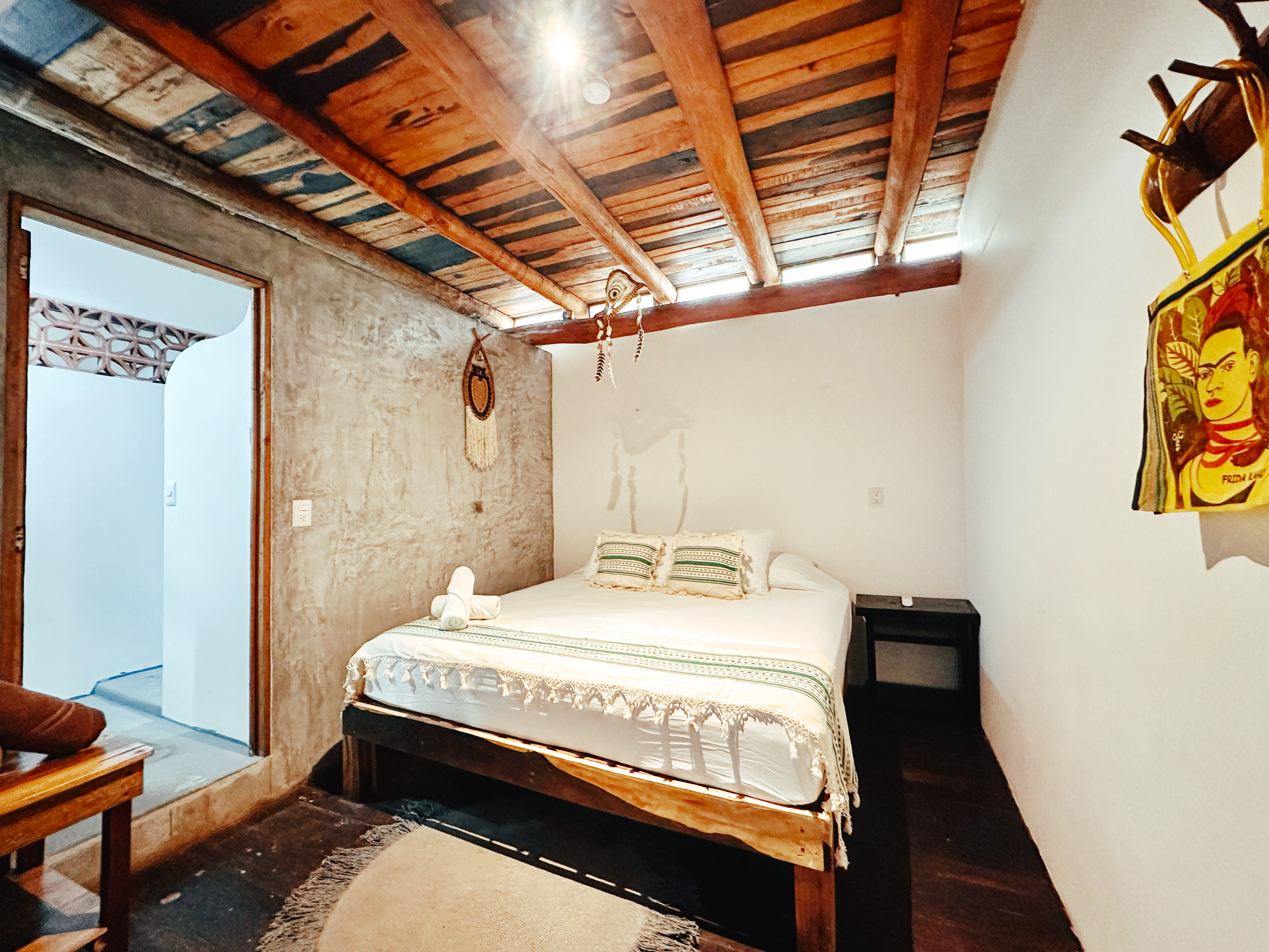 Small rustic bedroom with a bed, exposed wooden ceiling, white walls, and a colorful wall hanging
