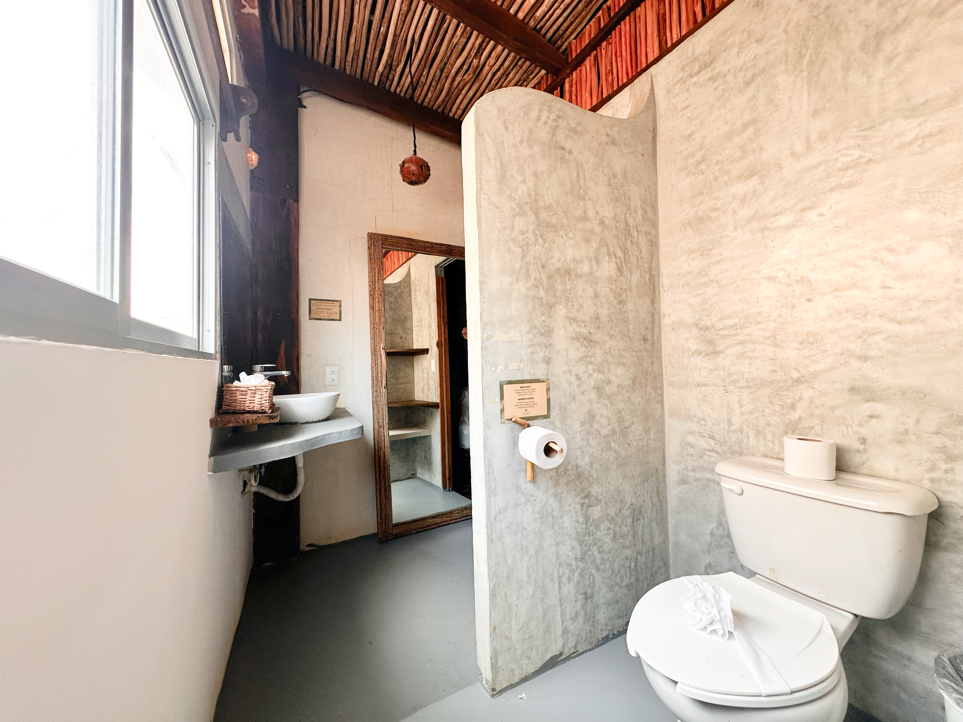 Small rustic bathroom with toilet, sink, mirror, and a window letting in daylight