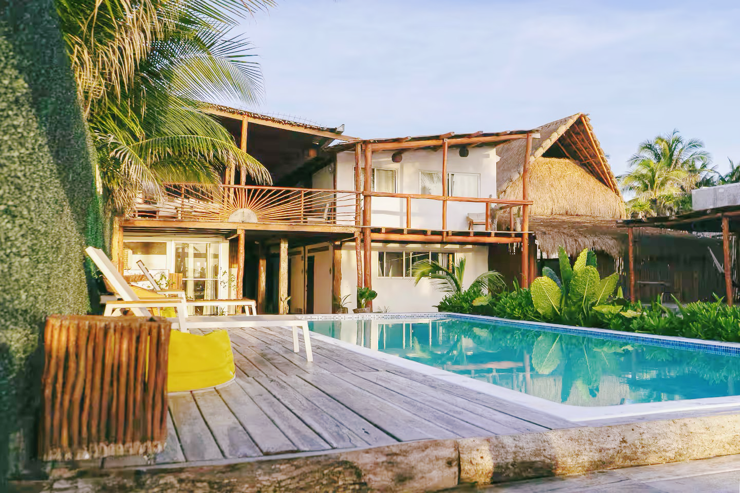 Tropical villa with a wooden deck and a turquoise swimming pool surrounded by palm trees.