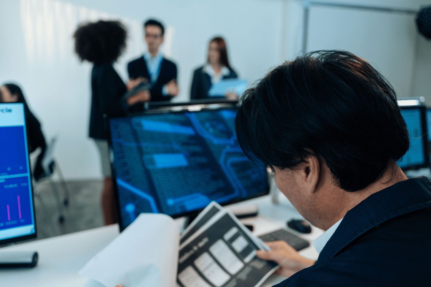 Person analyzing data on a laptop in a modern office, with coworkers and large monitors in the background