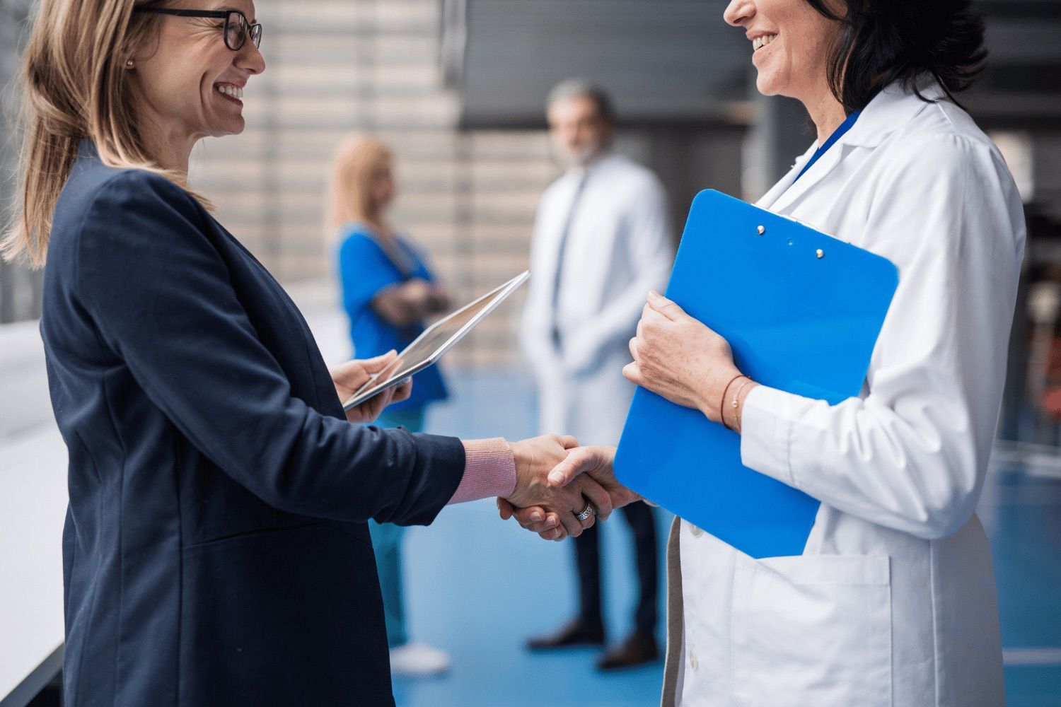 Woman in blue blazer shaking hands with doctor holding a blue clipboard in a hospital hallway