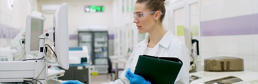 A lab worker in a white coat and safety glasses examines a computer, holding a clipboard in a medical lab.