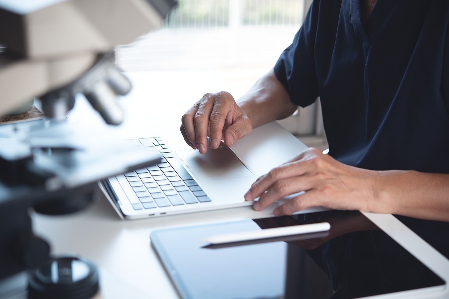 Hands typing on a laptop at a desk with a microscope nearby