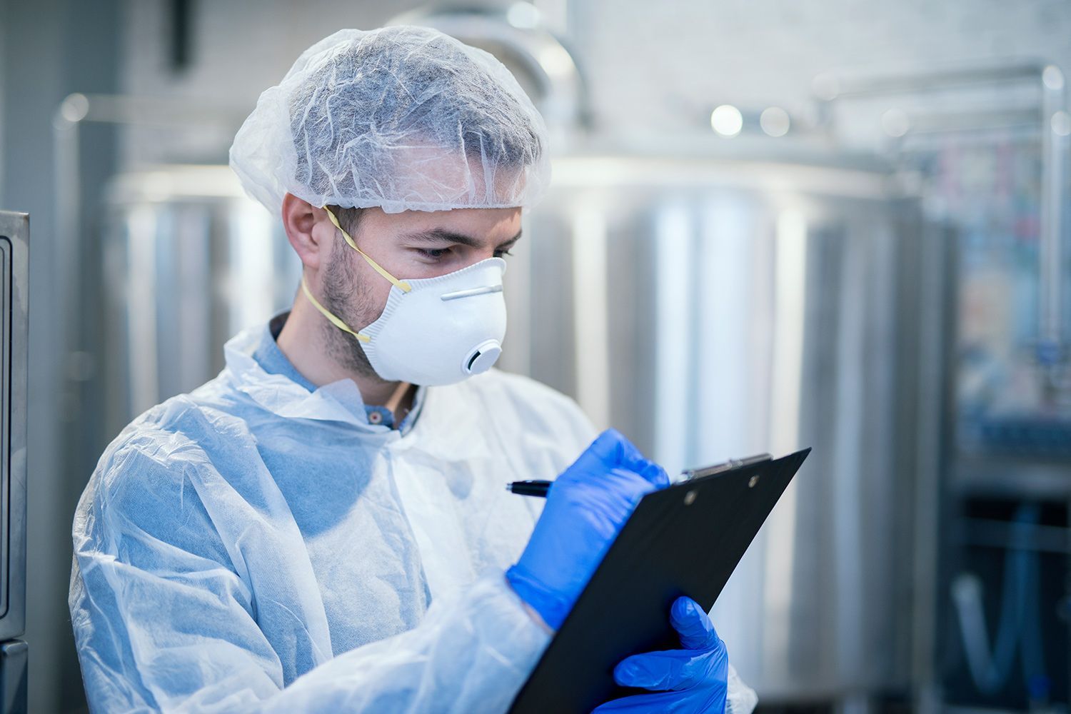 Lab technician in blue protective gear and mask writing on a clipboard in a clean industrial facility
