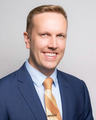 Man in blue suit, smiling at the camera, wearing a light blue shirt and gold-patterned tie.