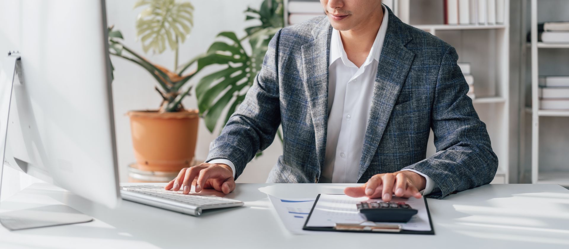 Man in blazer working at a desk with a computer, calculator, and documents.