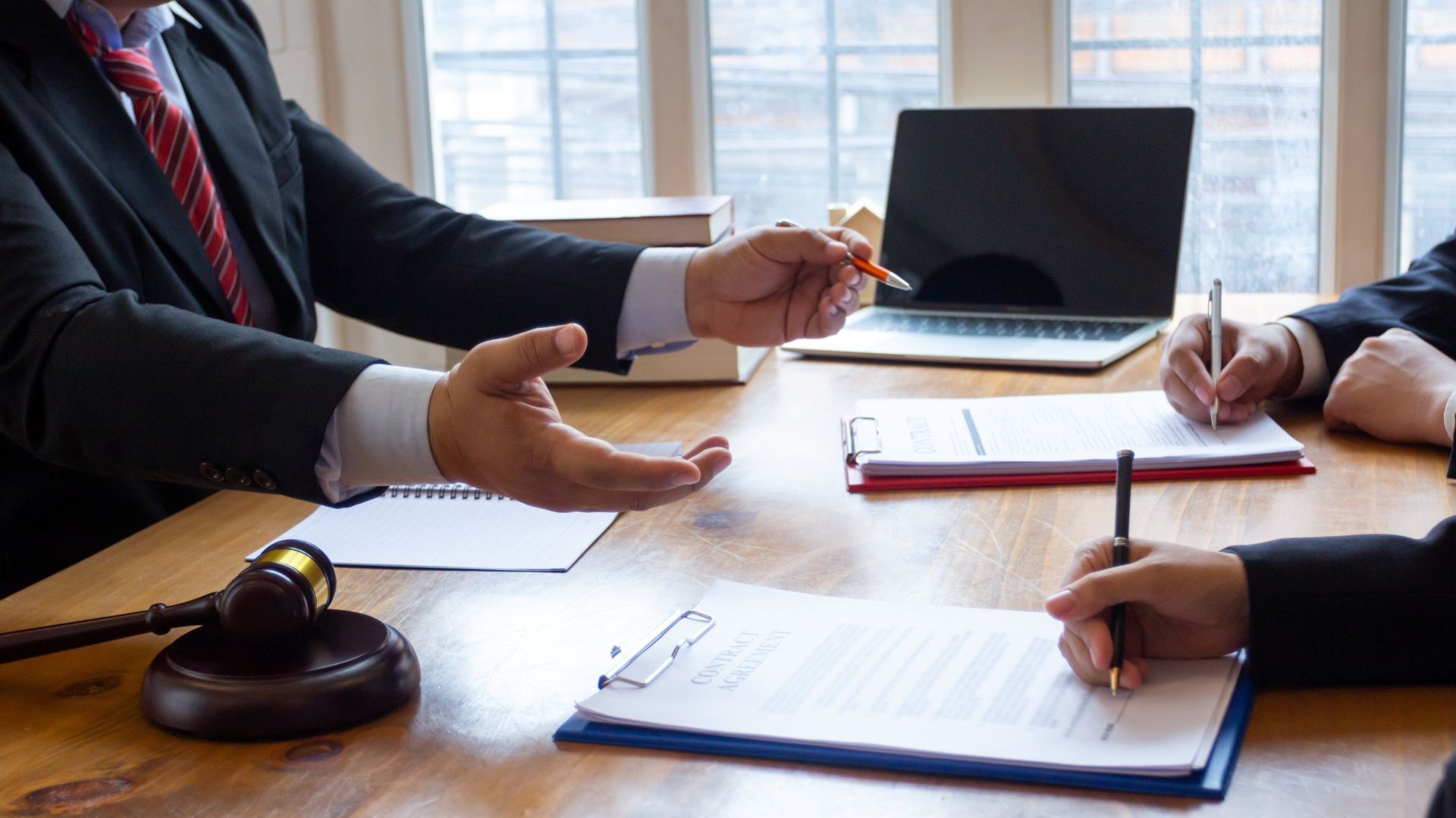 Lawyer gesturing to documents; two people take notes at a table with a gavel and laptop.