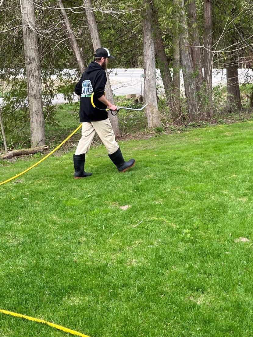 Man in black hoodie and boots spraying a lawn with a yellow hose.