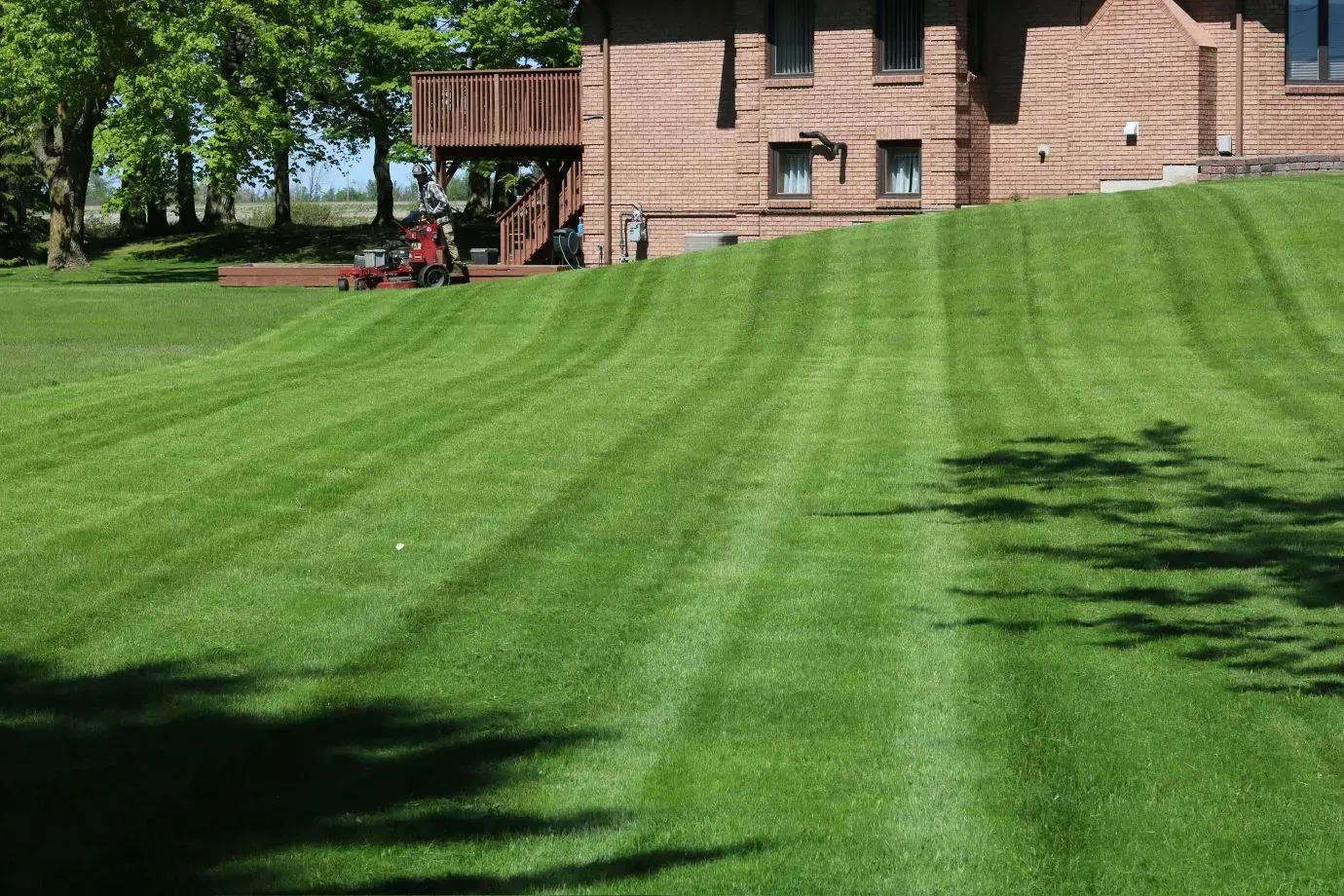 Lawn mowed with stripes, house with red brick facade, lawnmower on a hill. Green grass, blue sky.