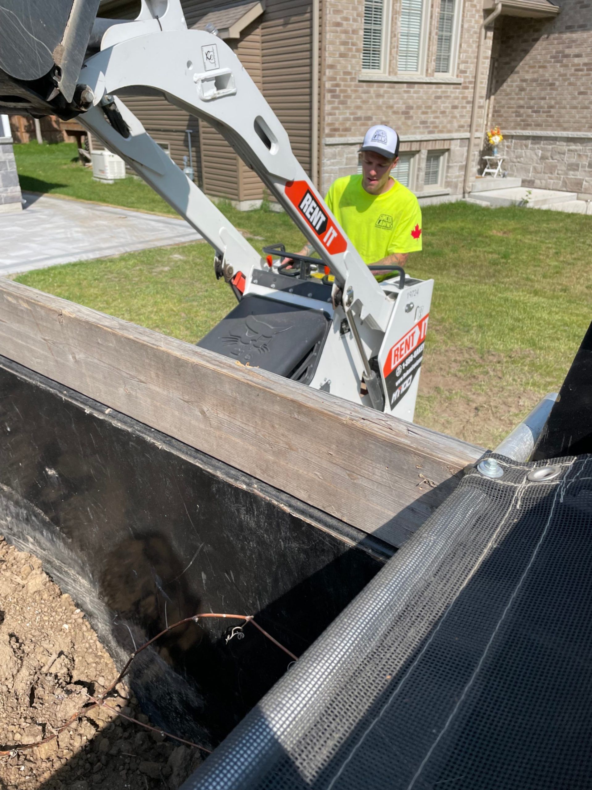A worker in a neon yellow shirt operates a Bobcat loader to move dirt into a black bin.