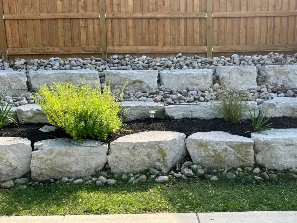 Stone retaining wall with greenery and gravel against a wooden fence.