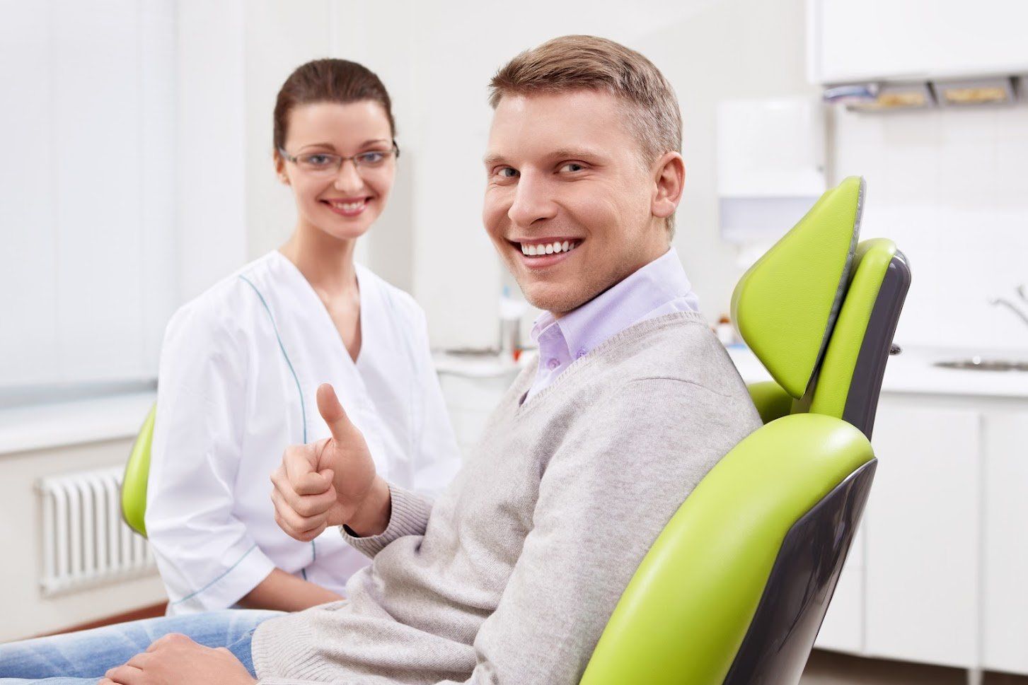 Man in dental chair gives thumbs up, smiling, with dentist smiling beside him in a white coat.