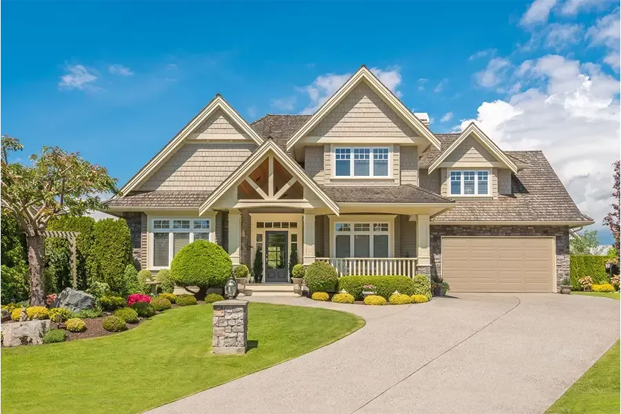 Suburban house with beige siding, gabled roof, manicured lawn, and a large driveway under a blue sky