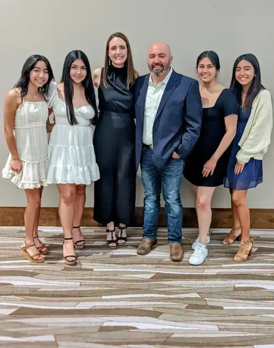 Group of six people standing indoors, smiling in formal and casual outfits against a plain wall.