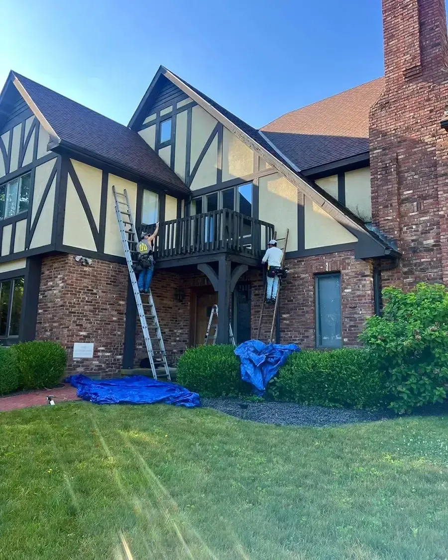Tudor-style house exterior with brick and timber framing; two ladders and blue tarps on the lawn during repairs