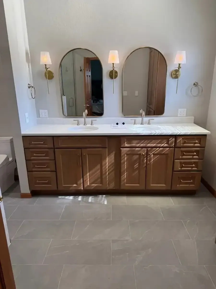 Empty bathroom with double vanity, round mirrors, wall sconces, and beige tile floor.