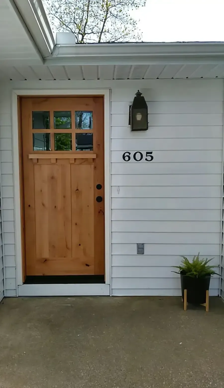 Front porch with wooden door, house number 605, black lantern, and potted plant beside white siding.