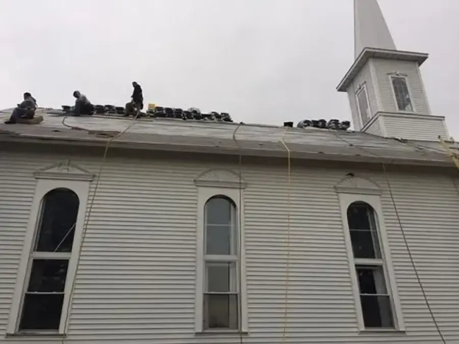 Workers on a white church roof, repairing shingles near the steeple and windows