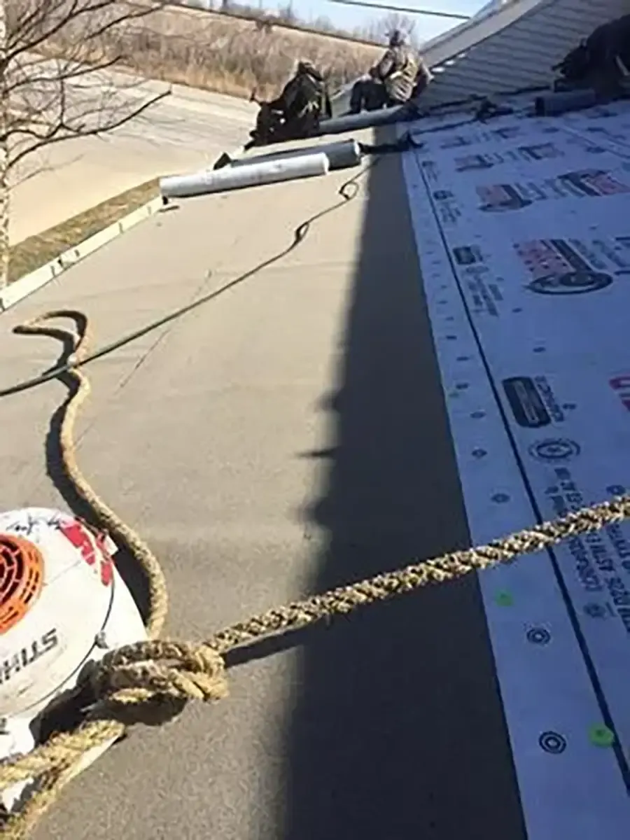 Workers installing roof underlayment on a sloped roof, with safety ropes and tools visible.