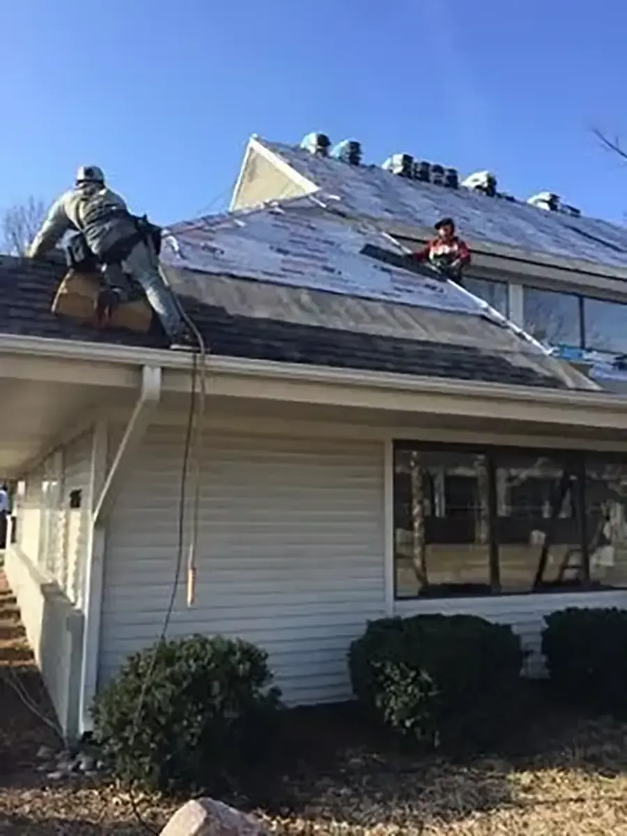 Two workers on a roof, one near the gutter and one on the slope, with shingles being replaced.