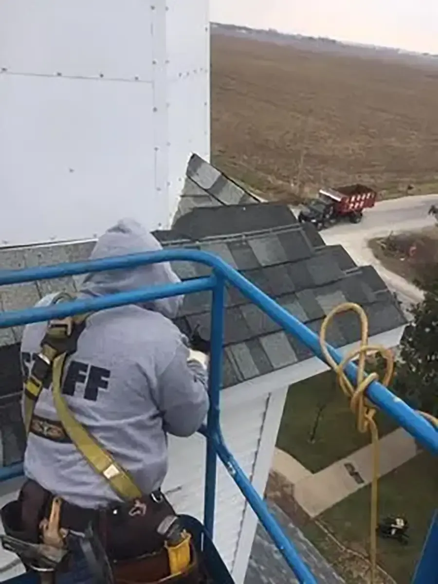 Worker in gray hoodie and safety harness on blue lift beside a white building roof.