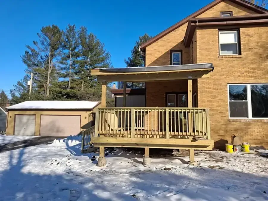 Snowy wooden house with porch and detached garage under a clear blue sky