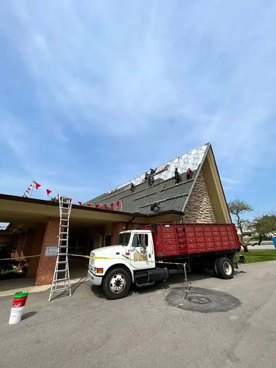 Red dump truck beside a building as workers repair the roof with ladders and shingles under a blue sky