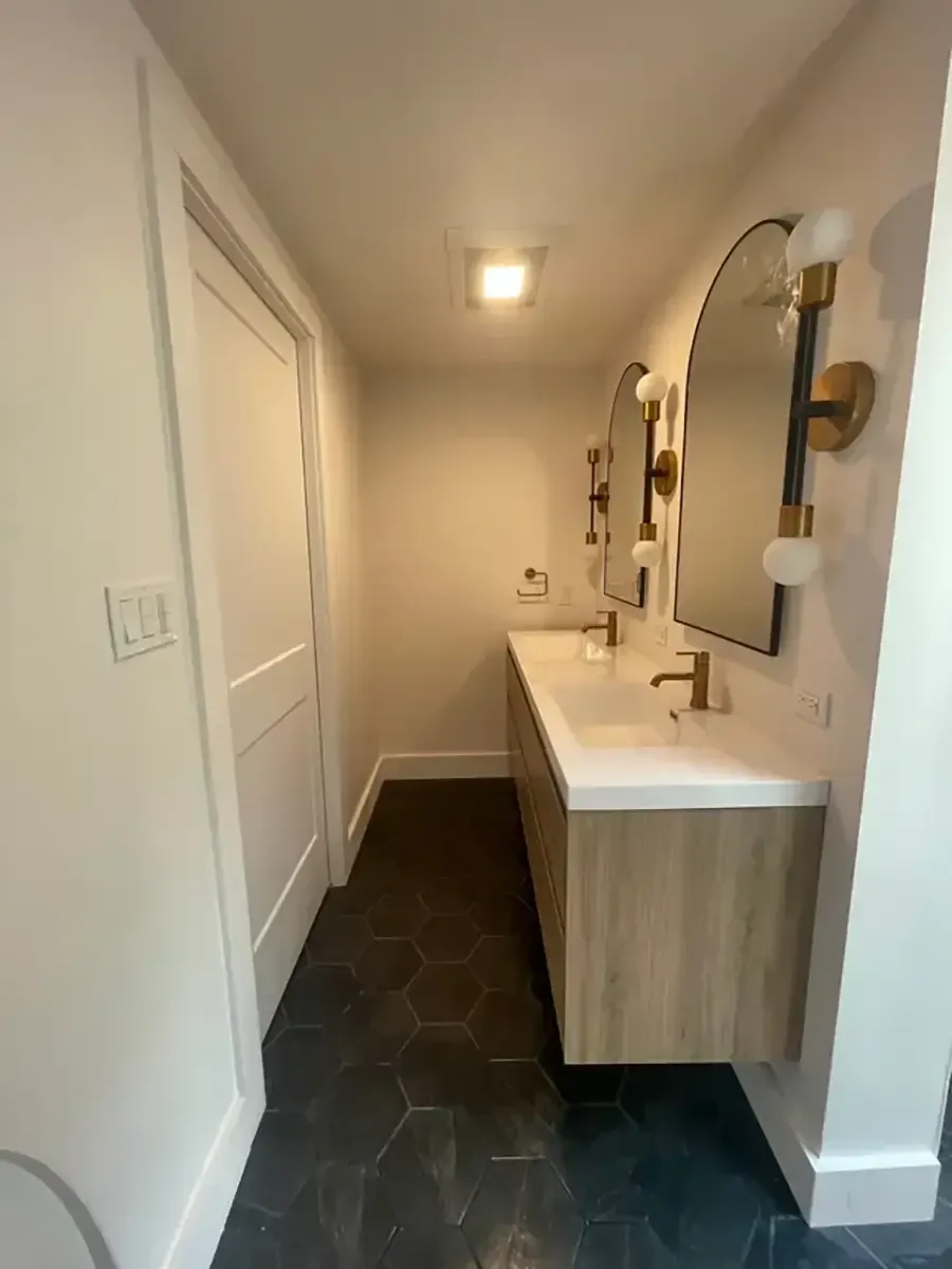 Narrow bathroom with double sink vanity, mirrors, and dark hex tile floor in a bright white hallway