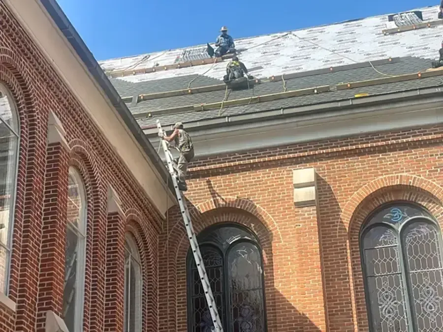 Worker on ladder repairing the roof of a brick building with arched windows.