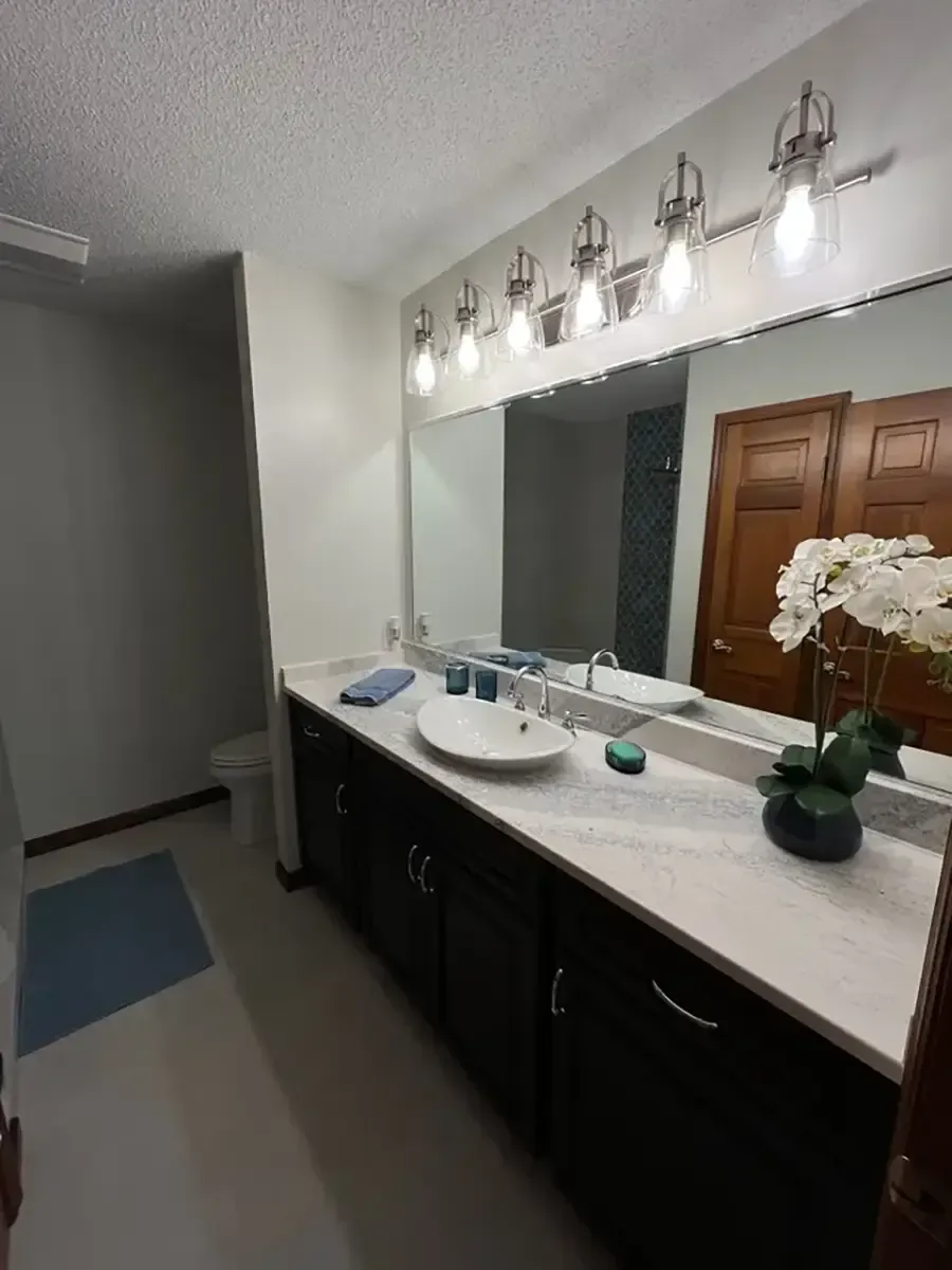 Bathroom vanity with large mirror, wall sconces, dark cabinets, and a white countertop with flowers and toiletries