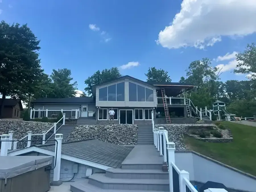 Lakeside house with large windows, stone retaining wall, and stairs leading down to a dock under a blue sky