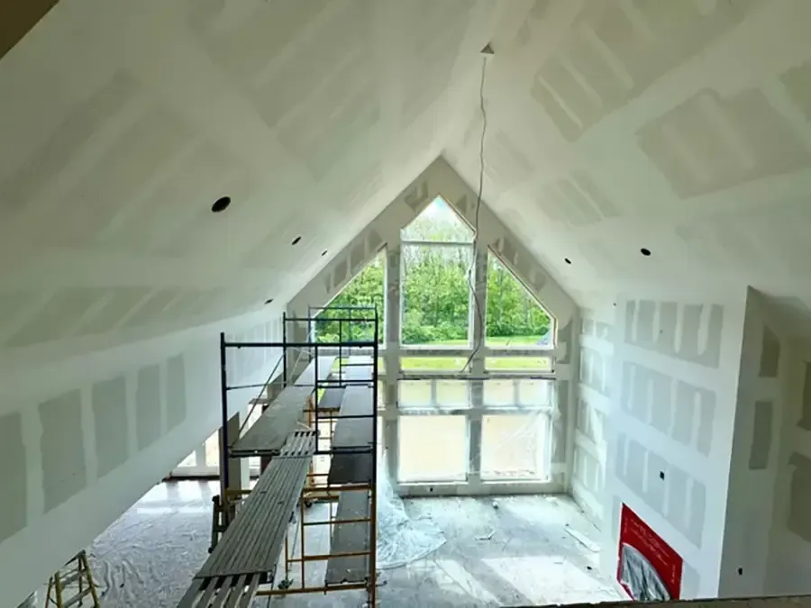 Attic room under construction with drywall, scaffolding, and a large triangular window at the end.