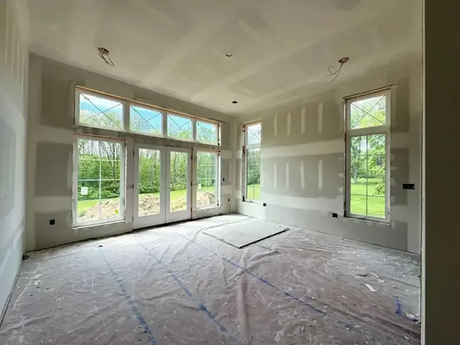 Empty sunlit room with large windows, unfinished drywall, and covered floor in a house under construction