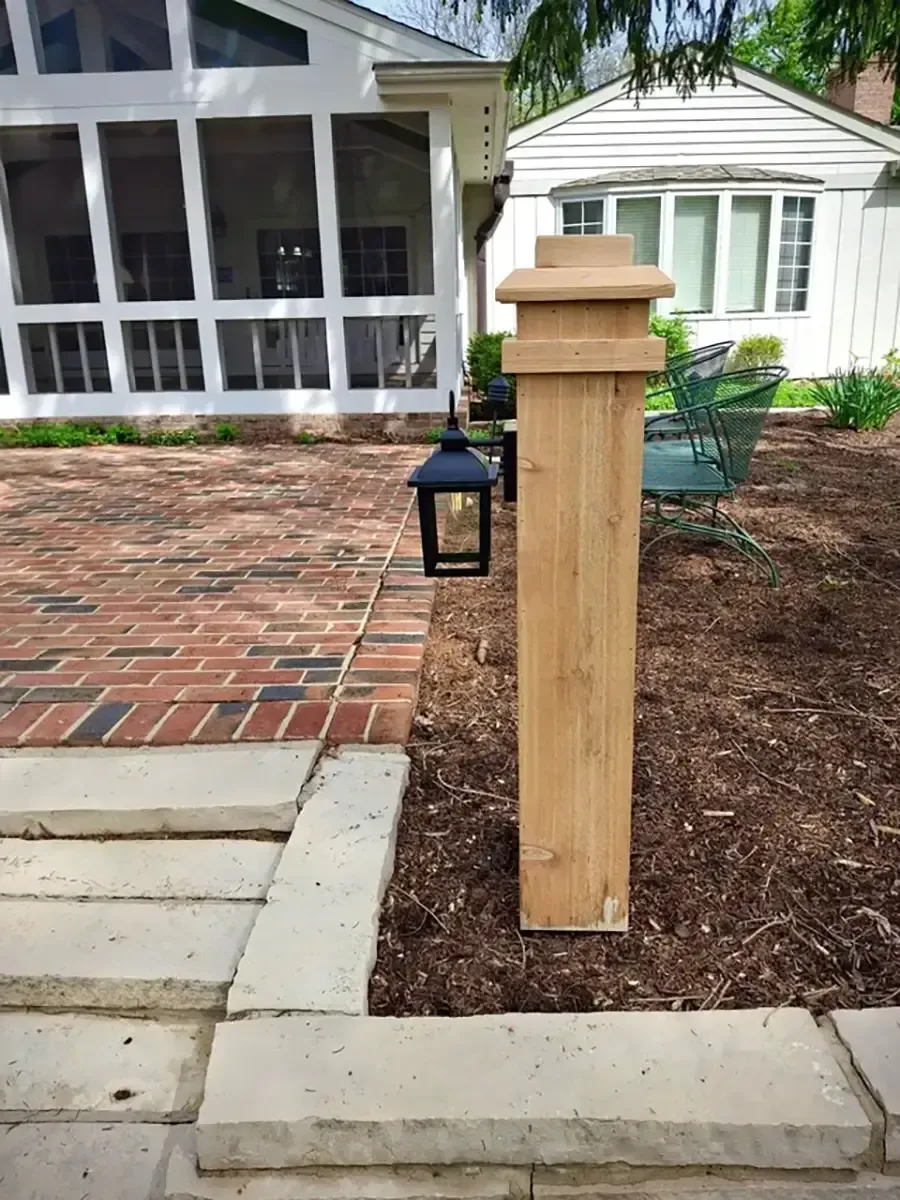 Wooden porch post beside a brick walkway, with a black lantern on a mulched garden bed.
