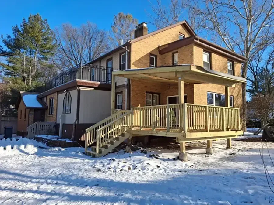 Snow-covered two-story house with a wooden porch and stairs under a clear blue sky