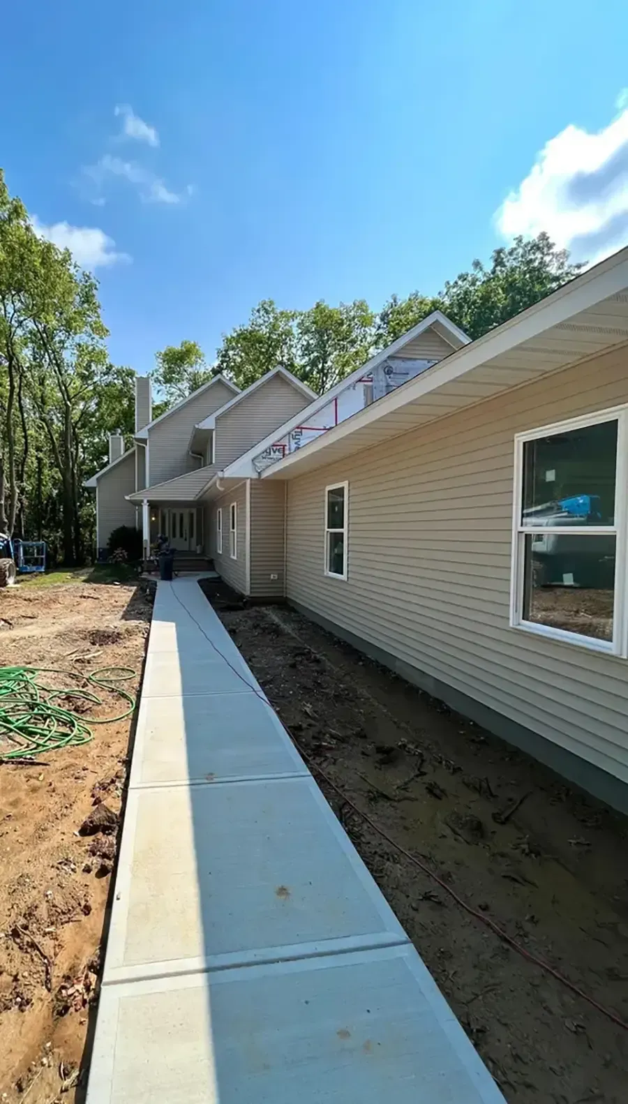 Concrete walkway beside a beige house under construction, with bare soil and trees under a blue sky