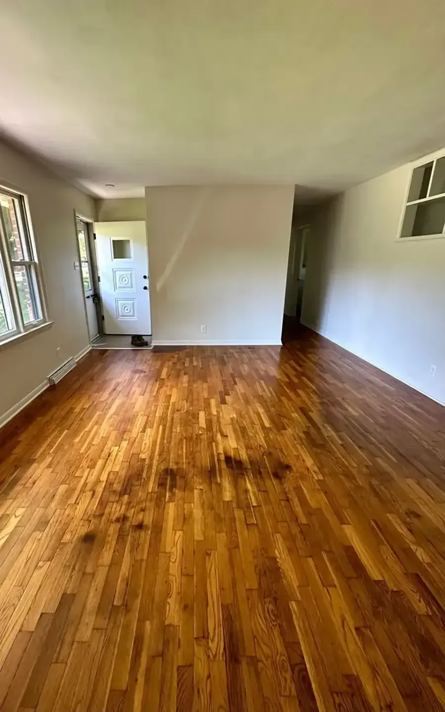 Empty living room with glossy hardwood floors, white walls, and windows along the left side.
